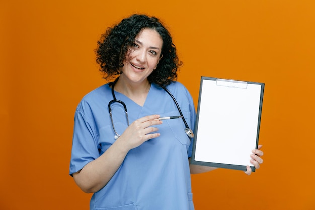 Улыбающаяся женщина-врач средних лет в униформе и со стетоскопом на шее смотрит в камеру, показывая буфер обмена, указывая на него ручкой, изолированной на оранжевом фоне