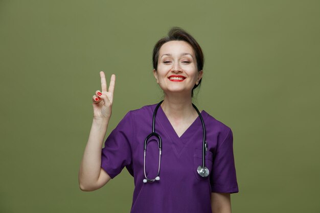 Улыбающаяся женщина-врач средних лет в форме и со стетоскопом на шее смотрит в камеру, показывающую знак мира на оливково-зеленом фоне