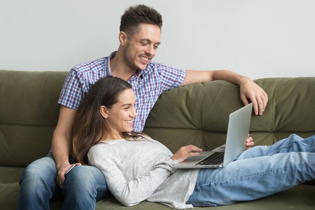 Smiling millennial couple enjoying using laptop relaxing on couch together