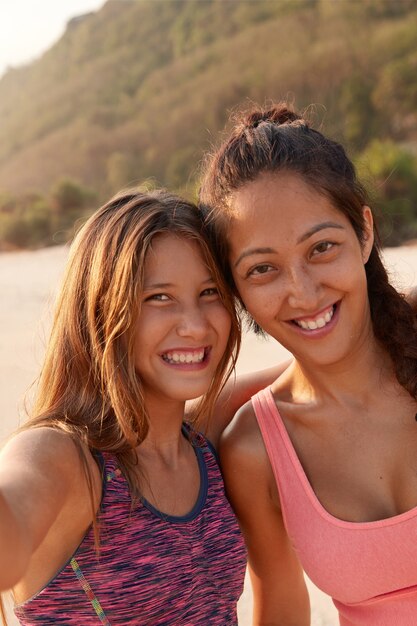 Smiling mixed race female friends pose near for making selfie, dressed in top