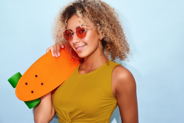 トレンディな夏の短いジーンズとスケートボードでポーズのサングラスで笑顔のモデルの女性