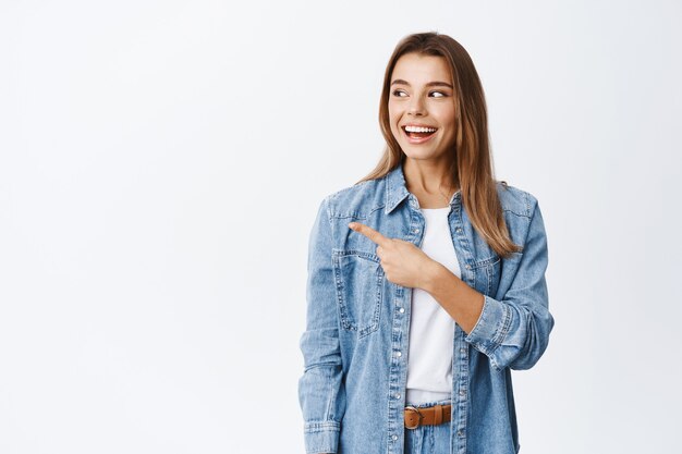 Smiling modern girl in casual clothes, pointing and staring left at logo with pleased face, checking out good deal on banner sideways, white wall