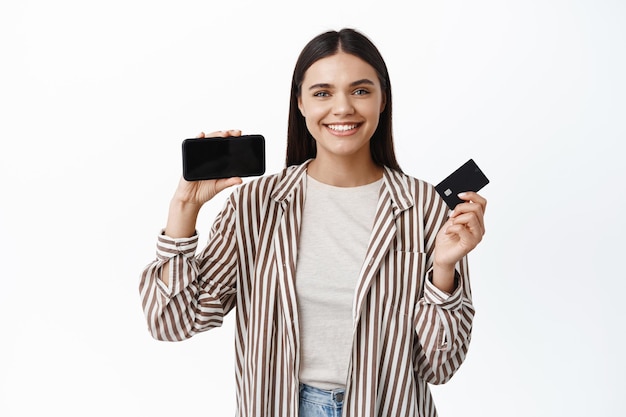 Smiling modern girl in stylish outfit, showing empty smartphone screen horizontally and plastic credit card, recommending an app, shopping online, white wall