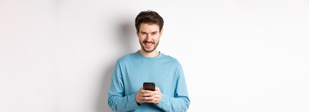 Smiling modern man using smartphone and looking pleased at camera guy in sweatshirt with mobile phon