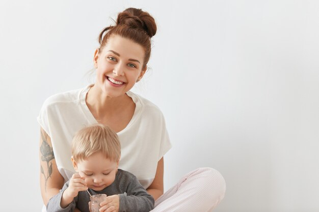 Smiling mother caring about her three-years old child on white wall. Lovely blond kid in grey shirt eating childhood nutrition from glass can with little spoon.