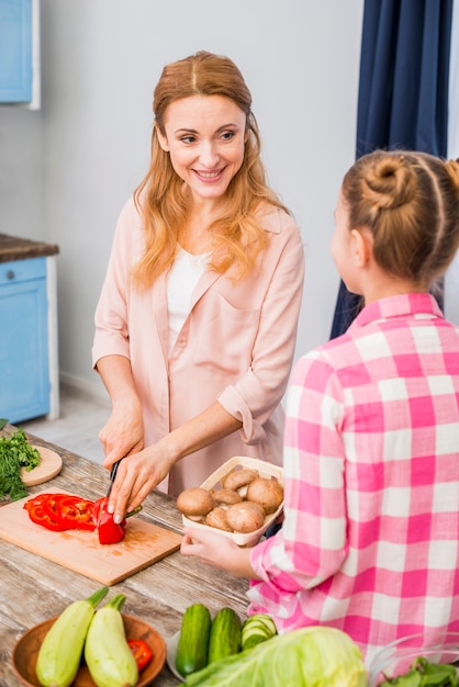 Smiling mother cutting the bell pepper with knife looking at her daughter holding mushroom basket