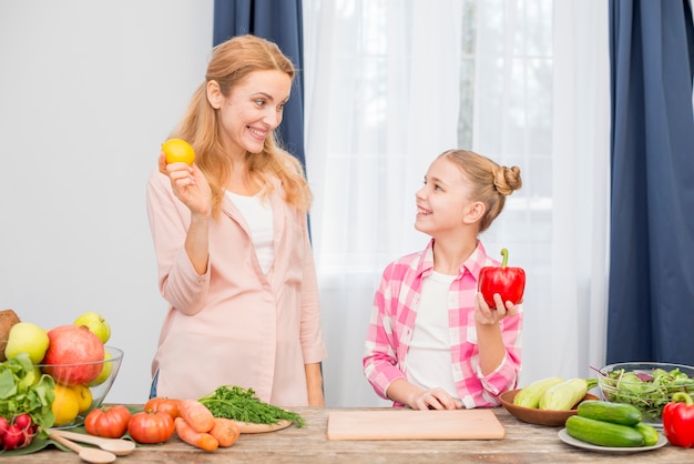 Smiling mother and daughter holding yellow lemon and red bell pepper in hand