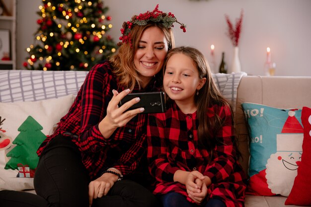 smiling mother and daughter looking at phone sitting on couch and enjoying christmas time at home