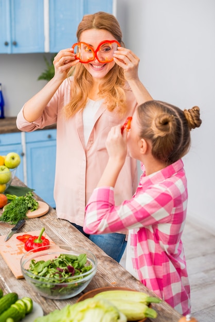 Madre e figlia sorridenti che guardano attraverso la fetta di peperone dolce rosso nella cucina