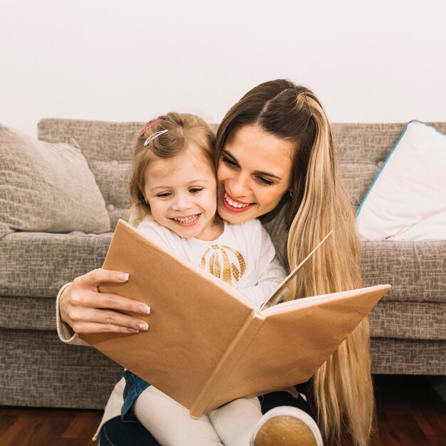 Smiling mother and daughter reading book near couch