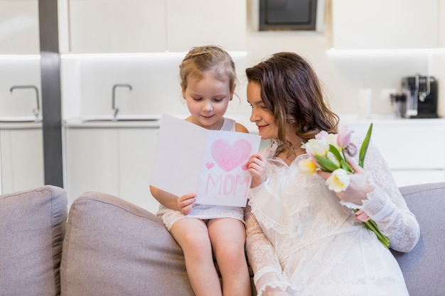 Smiling mother and daughter reading greeting card sitting on sofa with holding flower bouquet