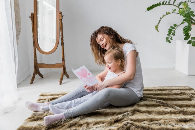 Smiling mother and daughter sitting on fluffy carpet looking greeting card in home