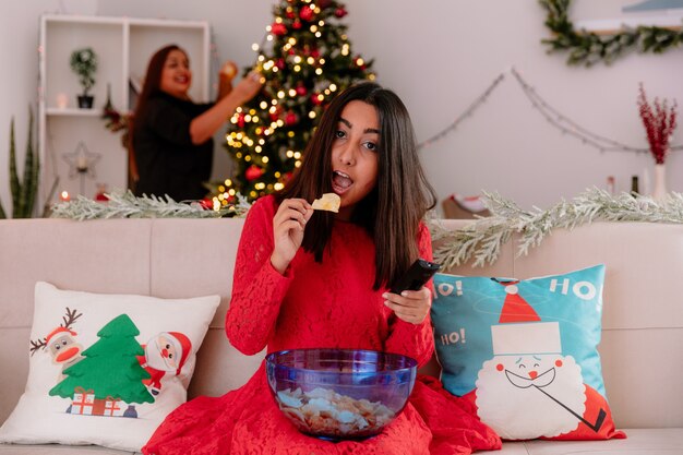 smiling mother decorates christmas tree and looks at her daughter eating bowl of chips sitting on couch enjoying christmas time at home