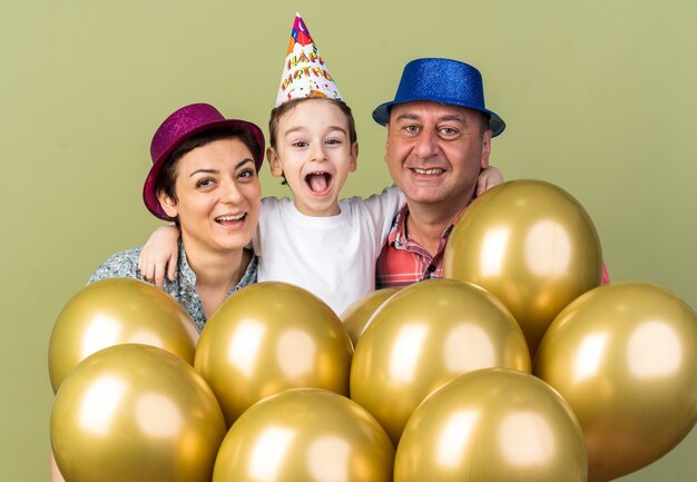 smiling mother and father wearing party hats standing with their son and holding helium balloons isolated on olive green wall with copy space