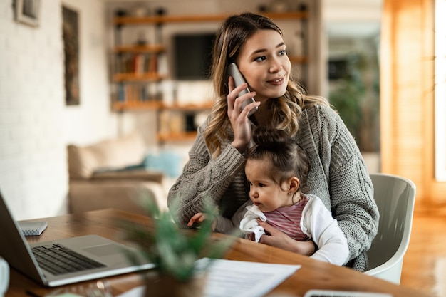 Улыбающаяся мать держит маленькую дочь на коленях во время разговора по телефону дома