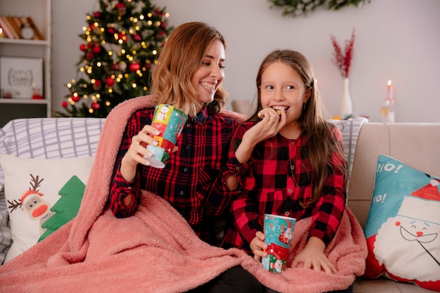 smiling mother holds paper cup and feeds daughter with biscuit sitting on couch covered with blanket and enjoying christmas time at home