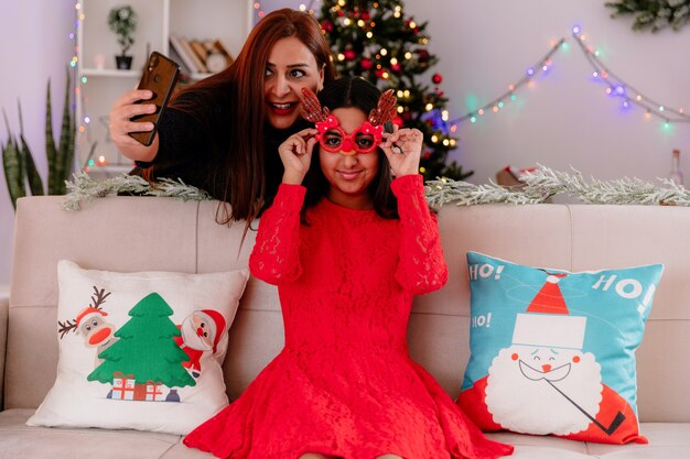 Улыбающаяся мать фотографирует свою дочь в оленьих очках, сидящую на диване и наслаждающуюся Рождеством дома