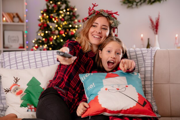 улыбающаяся мать с венком из падуба держит пульт от телевизора и очки, сидя на диване и наслаждаясь Рождеством с дочерью дома