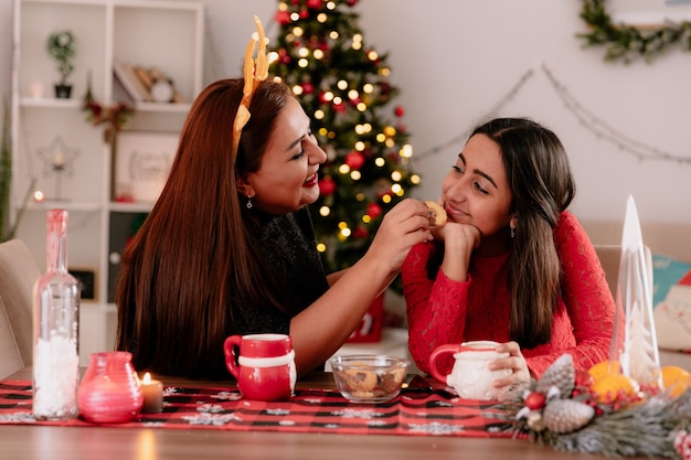 smiling mother with reindeer headband feeds her pleased daughter sitting at table enjoying the christmas time at home