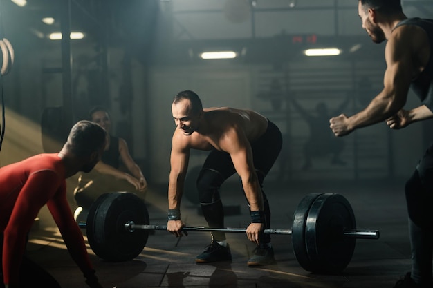 Улыбающийся мужчина мускулистого телосложения практикует становую тягу со штангой в клубе здоровья, пока его друзья подбадривают его