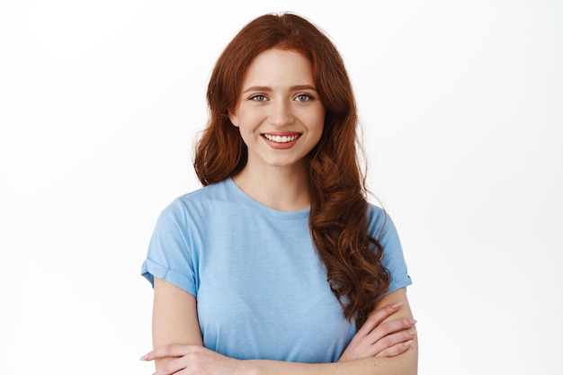 Smiling natural redhead girl, cross arms on chest, looks confident and determined at camera, professional woman in t-shirt, standing against white background