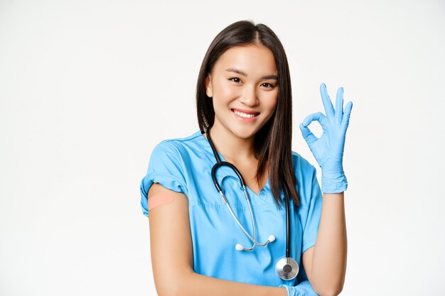 Улыбающаяся медсестра, азиатская женщина-врач в скрабах, показывающая знак «хорошо» и вакцинированная рука с медицинским пластырем, рекомендующая вакцинацию от covid-19, на белом фоне