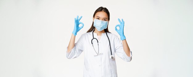 Smiling nurse female asian doctor in rubber gloves medical face mask shows okay ok signs stands agai