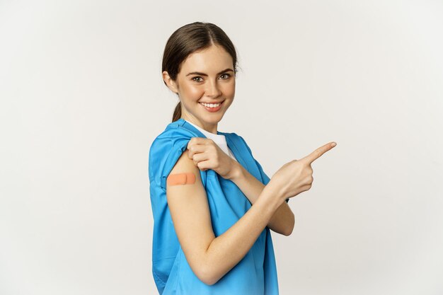 Улыбающаяся медсестра-медик в халате показывает пальцем вправо, показывая нашивку на плечевом ваку...
