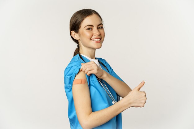 Smiling nurse, medical worker showing thumbs up and shoulder after vaccine, covid-19 vaccination campaign, standing over white background in scrubs.