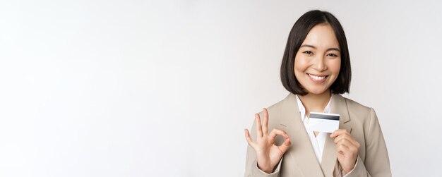 Smiling office clerk asian corporate woman showing credit card and okay sign recommending bank standing over white background in beige suit