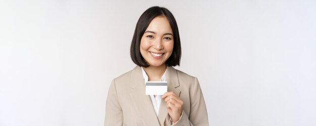 Smiling office clerk asian corporate woman showing credit card standing over white background in bei
