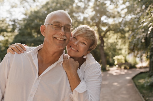 Smiling old man with grey hair and mustache in glasses and light shirt hugging with blonde woman in white clothes in park.