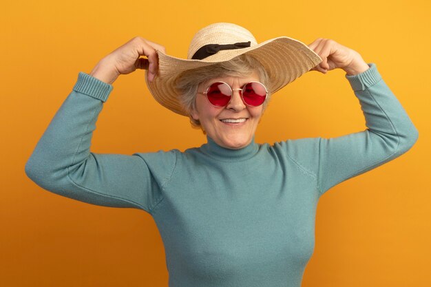 Улыбающаяся старуха в синей пляжной шляпе с водолазкой и солнцезащитными очками берет шляпу, изолированную на оранжевой стене