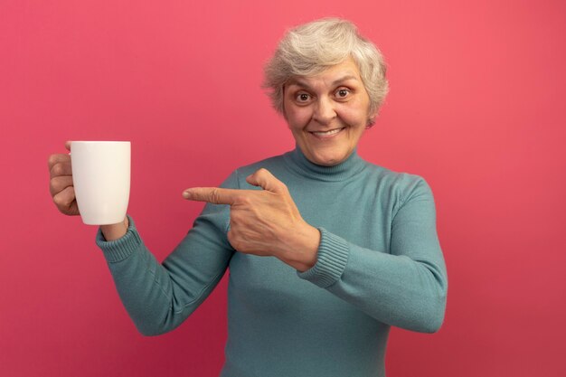 Smiling old woman wearing blue turtleneck sweater holding and pointing at cup of tea 