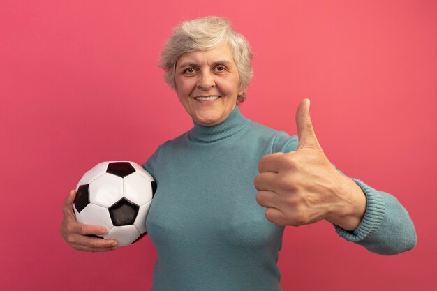 Smiling old woman wearing blue turtleneck sweater holding soccer ball looking at front showing thumb up isolated on pink wall