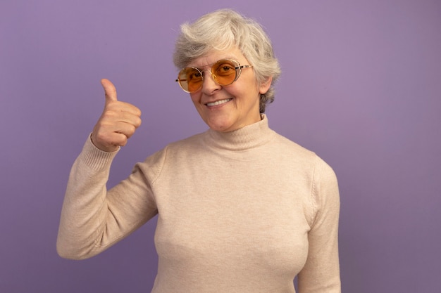 Улыбающаяся старуха в сливочном свитере с высоким воротом и солнцезащитных очках смотрит вперед, показывая большой палец вверх изолированно на фиолетовой стене