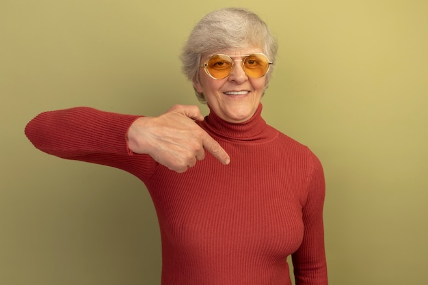 빨간 터틀넥 스웨터와 선글라스를 끼고 웃고 있는 노부인이 복사공간이 있는 올리브 녹색 벽에 고립된 정면을 바라보고 있다