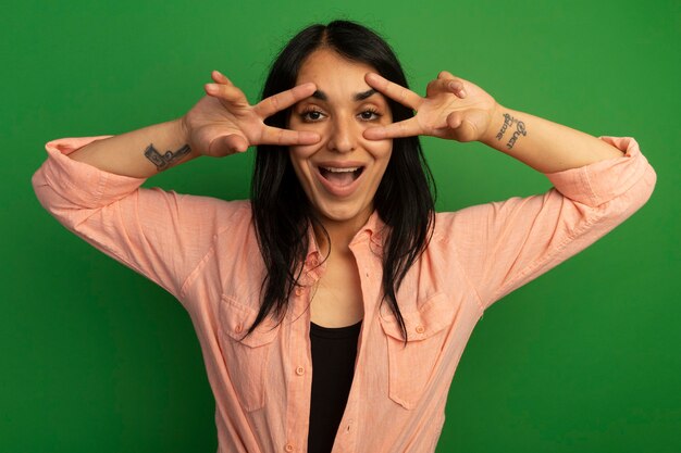 Smiling oung beautiful girl wearing pink t-shirt showing peace gesture isolated on green wall