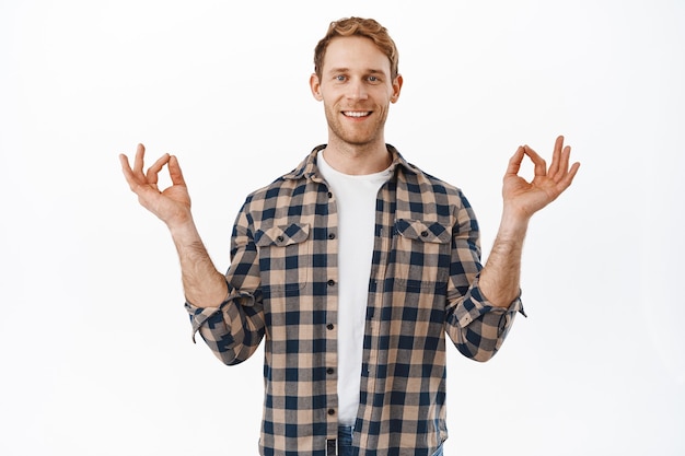 Smiling patient and relaxed redhead adult man, meditating, feel peaceful, breathing and resting on yoga, holding hands in lotus nirvana pose, white wall