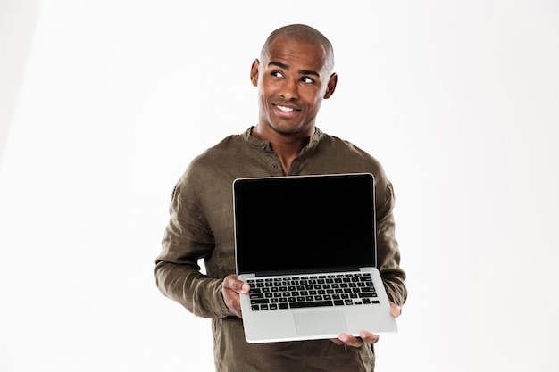 Smiling pensive african man showing blank laptop computer screen and looking up