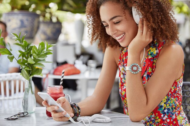 Smiling pleased dark skinned female model spends leisure time at outdoor cafe, uses modern technologies for listening favourite music via headphones