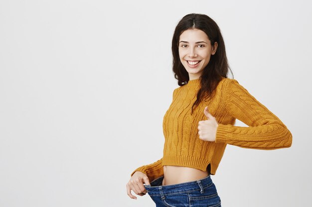 Smiling pleased girl lost weight, showing loose jeans and thumbs-up