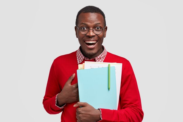 Smiling pleased male scientific worker keeps hand on chest, carries papers and notebook with pen, cant believe in his success, has joyful facial expression, wears round glasses