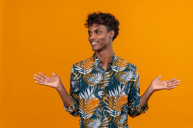 Free photo smiling pleased young handsome dark-skinned man with curly hair in leaves printed shirt opening hands on an orange background