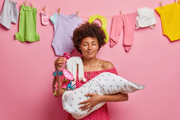 Smiling pleased young mother holds newborn child on hands, nurses little baby with mobile toy, enjoys calmness while newborn sleeping, poses against pink wall with kids clothes on rope