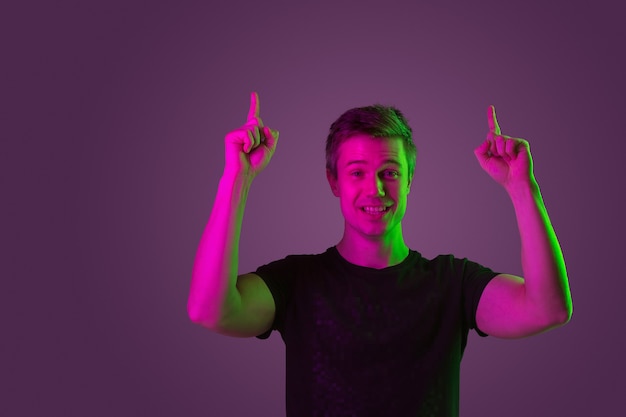 Smiling, pointing up. Copyspace. Caucasian man's portrait on purple studio background in neon light. Beautiful male model in black shirt. Concept of human emotions, facial expression, sales, ad.