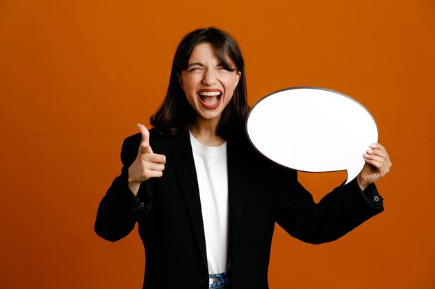 オレンジ色の背景に分離された黒いジャケットを着て吹き出し若い美しい女性を保持しているカメラで笑顔のポイント
