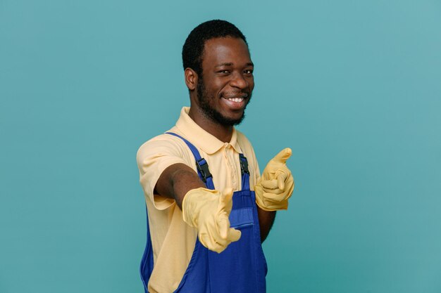 Smiling points at camera young africanamerican cleaner male in uniform with gloves isolated on blue background