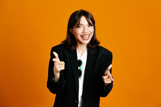 Smiling points at camera young beautiful female wearing black jacket isolated on orange background