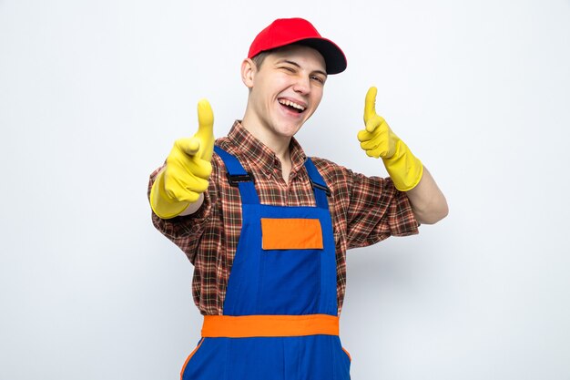 Smiling points at front young cleaning guy wearing uniform and cap with gloves